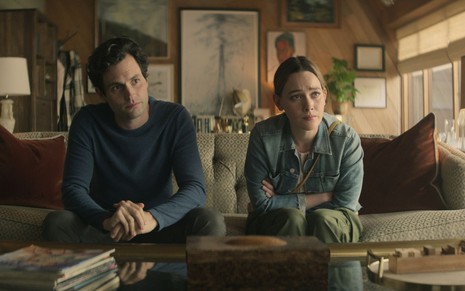 Penn Badgley e Victoria Pedretti sentados lado a lado em um sofá em cena da 3ª temporada de You