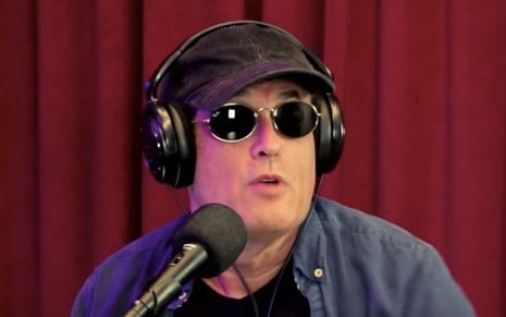 Sergio Mallandro de óculos escuros, fone de ouvido, em frente a microfone, com cortina vermelha ao fundo