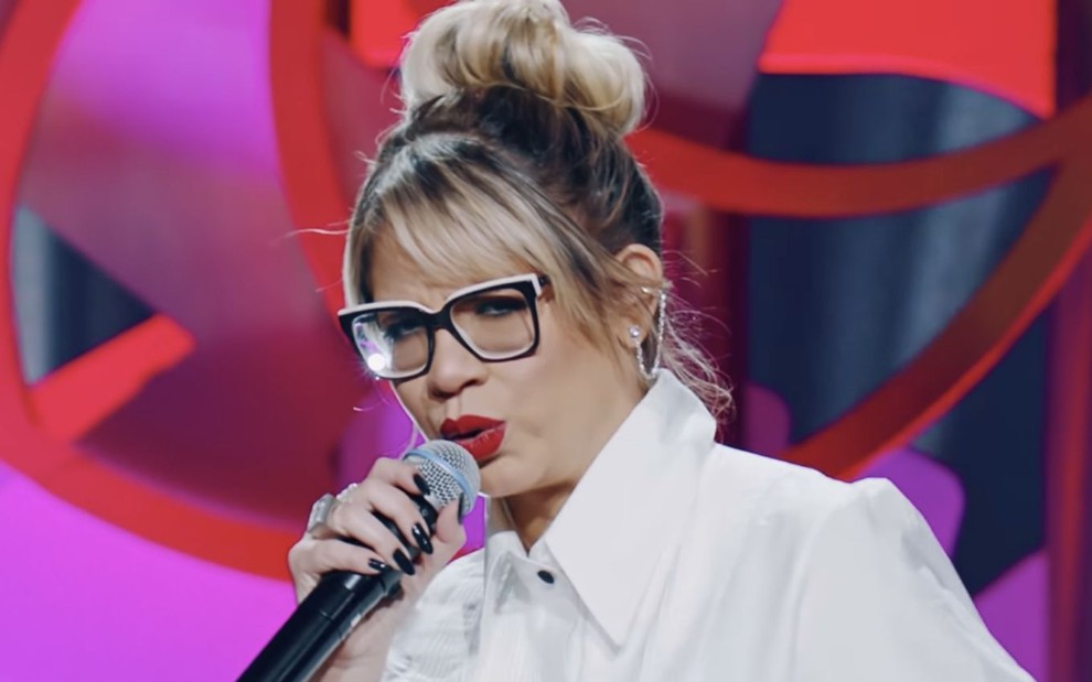 A cantora sertaneja Marília Mendonça no clipe de Todo Mundo Menos Você; ela aparece segurando um microfone, com o cabelo preso e vestindo camisa branca