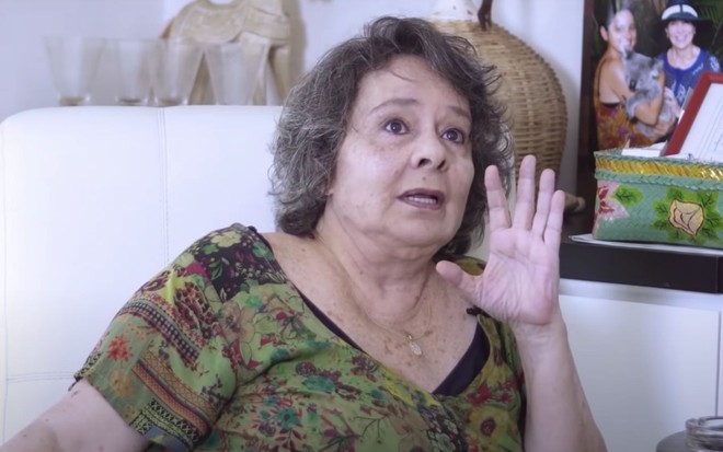 Débora Duarte em entrevista para o canal do YouTube de Paloma Duarte: atriz olha para cima, estica a palma da mão esquerda e está sentada