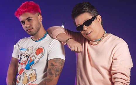 Zé Felipe veste blusa branca com desenho colorido, está com o cabelo rosa e aparece ao lado de DJ Ivis; o DJ usa óculos escuros, blusa rosa e está na frente de um fundo roxo