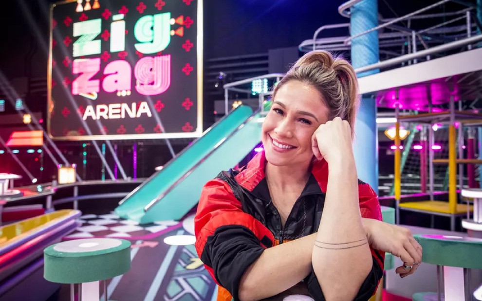 Fernanda Gentil usa uma camisa jeans e está com um microfone preto na mão no cenário do Zig Zag Arena