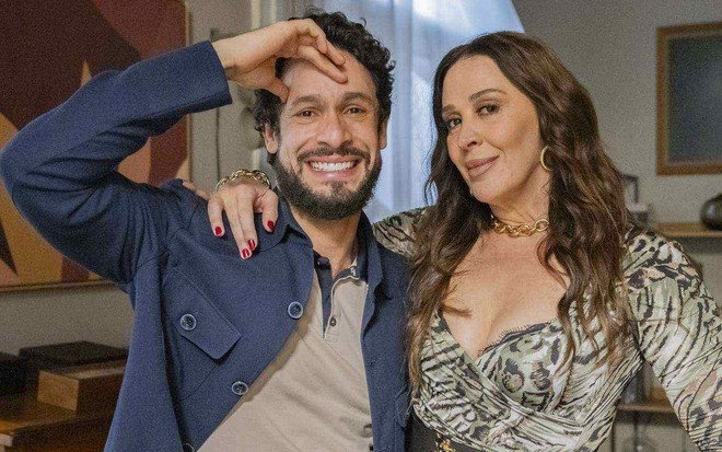 Rainer Cadete está com a mão na testa e faz um sorriso tenso abraçado com Claudia Raia em cenário da novela Terra e Paixão