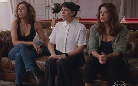 Em cena de Elas por Elas, Thalita Carauta, Karine Telles e Mariana Santos estão sentadas lado a lado em um sofá, preocupadas