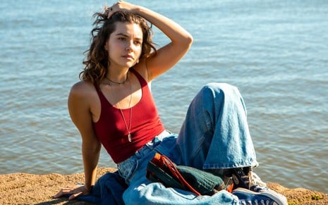 Alanis Guillen usa calça jeans e blusa vermelha e está sentada em uma pedra, com o mar ao fundo