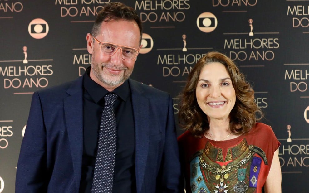 Alessandro Marson e Thereza Falcão posam sorridentes em frente a um painel do Melhores do Ano de 2023