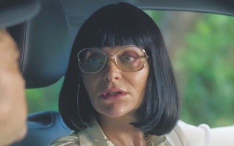 Alexandra Richter com expressão séria, cabelo curto e usando óculos de grau dentro de um carro