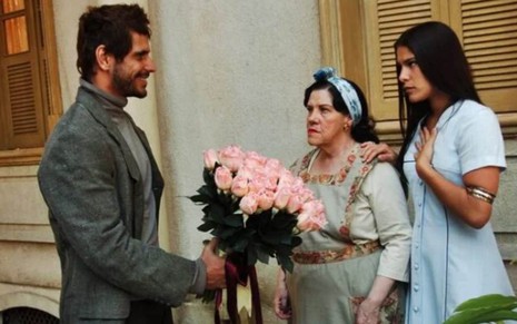 O ator Alexandre Barillari segurando um buquê de flores em frente às atrizes Neusa Maria Faro e Priscila Fantin, sérias, em cena de Alma Gêmea