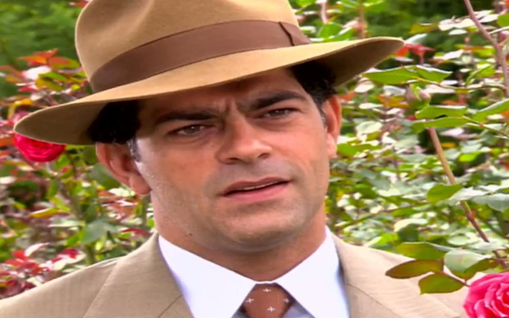 O ator Eduardo Moscovis de chapéu, em cena em roseiral, com expressão séria em Alma Gêmea