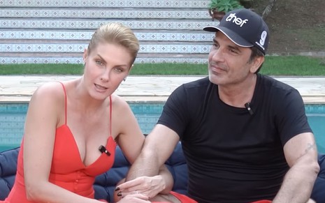 Ana Hickmann e Edu Guedes estão sentados na frente de uma piscina, de mãos dadas; eles encaram a câmera
