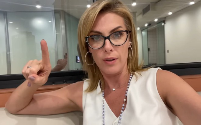 A apresentadora Ana Hickmann levanta o dedo indicador da mão direita, com uma roupa branca, de óculos, em seu camarim na Record