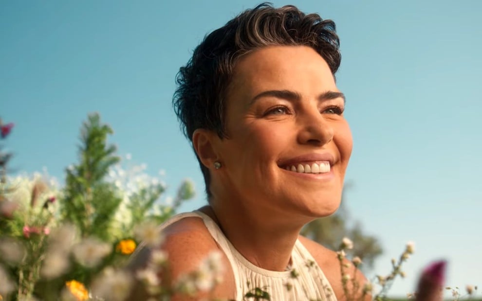 Ana Paula Arósio sorri em um campo florido em comercial para marca de suplementos de uma rede de farmácias
