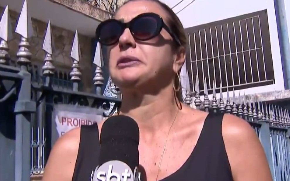 Andreia Andrade usa uma regata preta e óculos escuro; ela parece abalada ao dar entrevista ao SBT