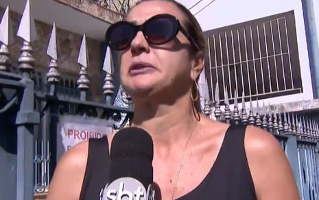 Andreia Andrade usa uma regata preta e óculos escuro; ela parece abalada ao dar entrevista ao SBT