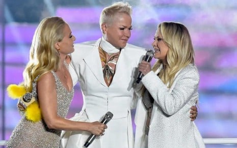 Angélica e Xuxa observam Eliana falando ao microfone no palco do Criança Esperança 2023