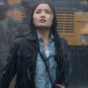 Anna Sawai caracterizada como Cate Randa, em frente a um ônibus escolar, tomando chuva e olhando para cima