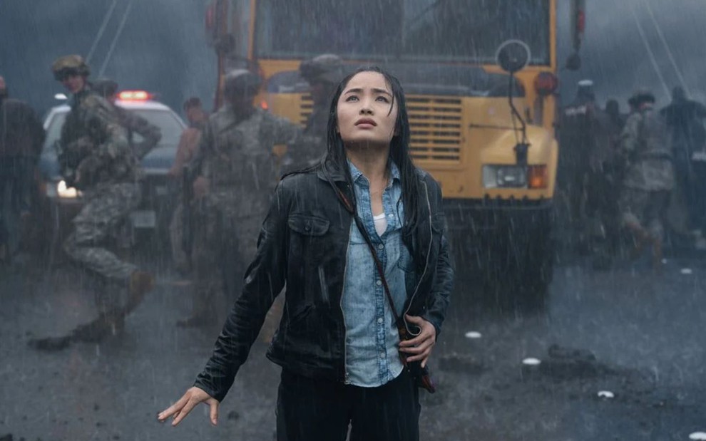 Anna Sawai caracterizada como Cate Randa, em frente a um ônibus escolar, tomando chuva e olhando para cima