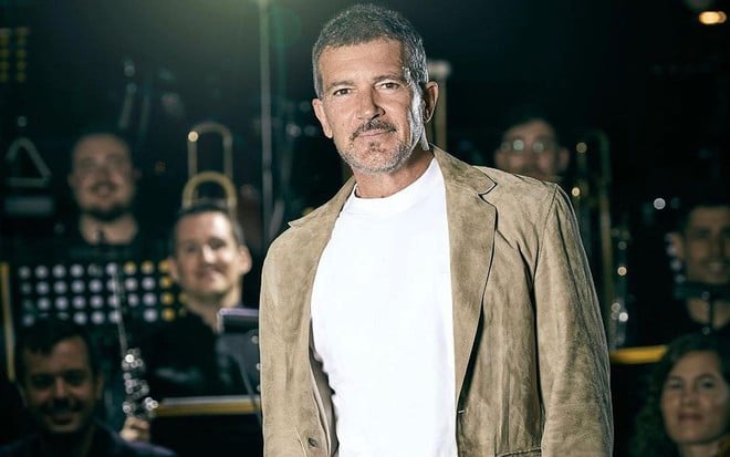 Antonio Banderas usa blusa branca e blazer marrom; ele parece estar em cima de um palco com pessoas atrás