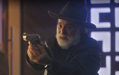 Em cena de Terra e Paixão, Tony Ramos usa chapéu e está com a arma apontada para alguém