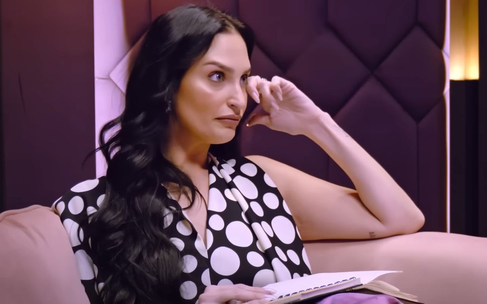 Ariela Carasso está sentada em sofá e parece enxugar uma lágrima discretamente