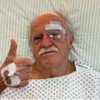 Ary Fontoura está deitado em uma cama de hospital, com um curativo no olho esquerdo, e faz sinal positivo com os dedos
