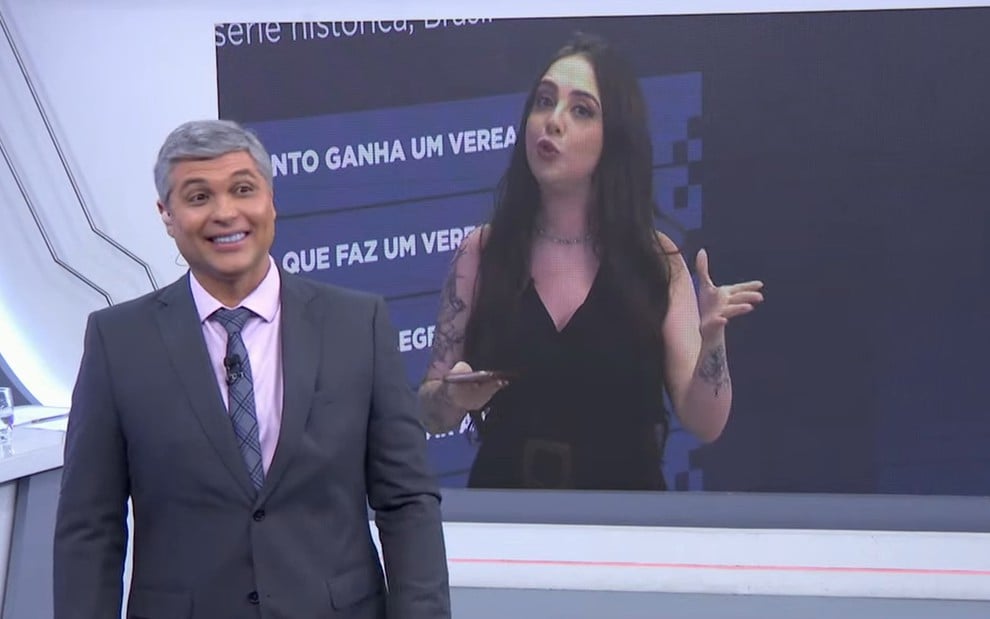 Joel Datena força sorriso no cenário do Brasil Urgente; a repórter Carine Roma está no telão do programa