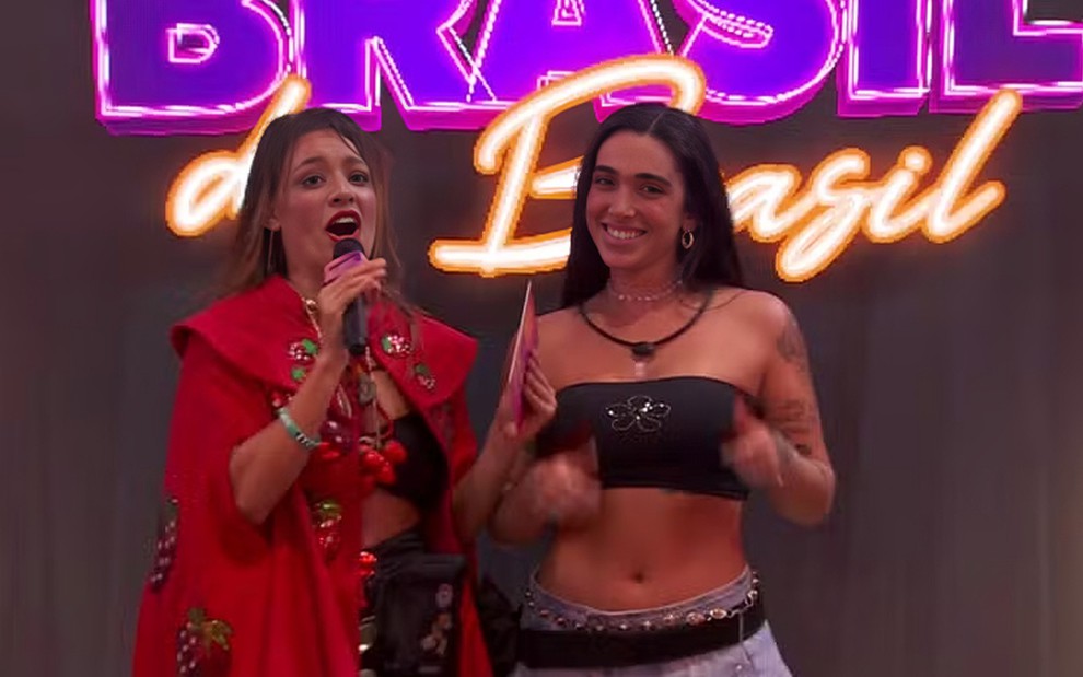 Com um sobretudo vermelho, Beatriz Reis faz careta enquanto segura microfone; Giovanna Lima sorri para a câmera