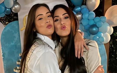 Deniziane Ferreira abraça a irmã gêmea, Enny Ferreira, em uma festa de aniversário; com balões azuis e prata ao fundo