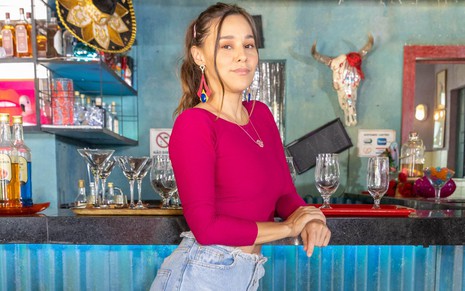 Thati Lopes posa para foto no cenário do bar de Terra e Paixão usando uma blusa rosa de manga comprida e uma saia jeans