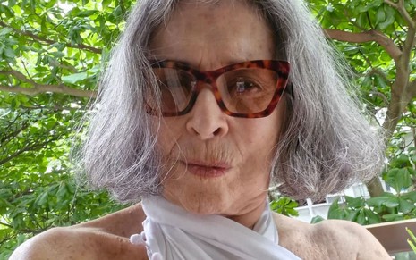 Betty Faria usa um óculos com animal print e uma blusa frente única branca; ela faz um bico para a câmera