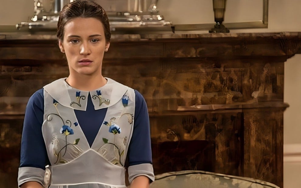 Em cena de Êta Mundo Bom!, Bianca Bin usa uniforme de doméstica e está parada, olhando para frente