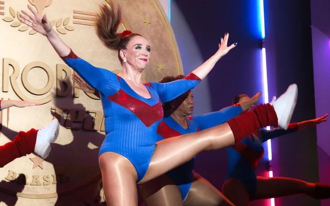 De maiô azul e vermelho, Marisa Orth está com a perna levantada fazendo ginástica