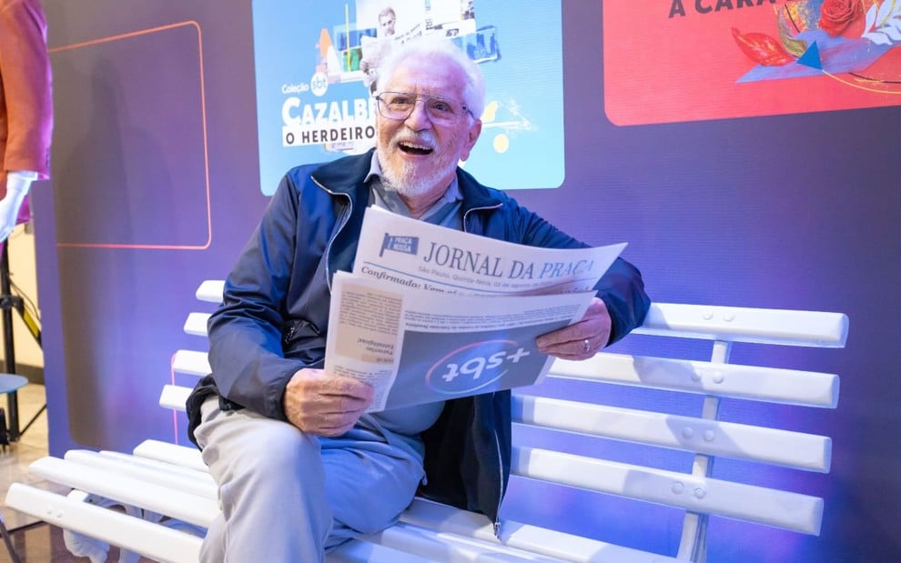 Carlos Alberto de Nóbrega está sentado no banco do A Praça É Nossa, fingindo ler uma edição do jornal que noticia o +SBT; ele sorri