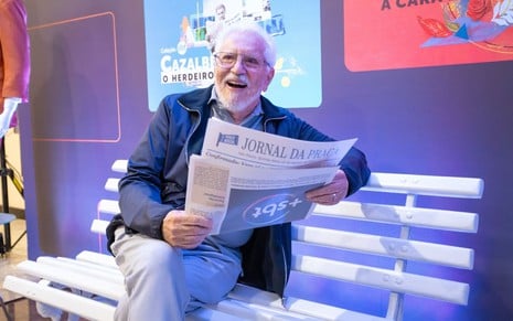 Carlos Alberto de Nóbrega está sentado no banco do A Praça É Nossa, fingindo ler uma edição do jornal que noticia o +SBT; ele sorri