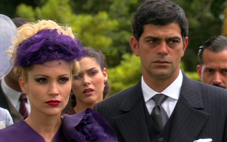 Os atores Flávia Alessandra e Eduardo Moscovis sérios, ela de chapéu roxo, ele de terno e gravata, em cena de Alma Gêmea