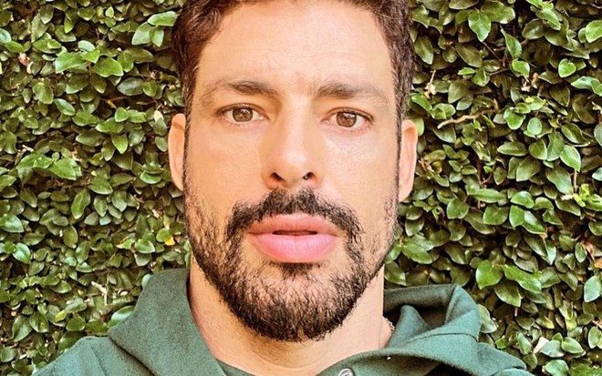 Cauã Reymond usa um moletom verde e está parado na frente de um canteiro de plantas