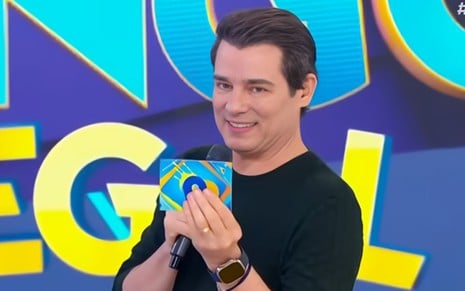 A imagem mostra um homem sorridente, de cabelo escuro, segurando um microfone e um cartão colorido em um estúdio de TV com um fundo azul vibrante.