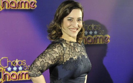 A atriz Simone Gutierrez sorrindo, posando para foto em frente a background com o logo da novela Cheias de Charme