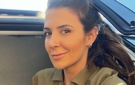 Cris Amaral usa um macacão verde e dá um leve sorriso para a câmera