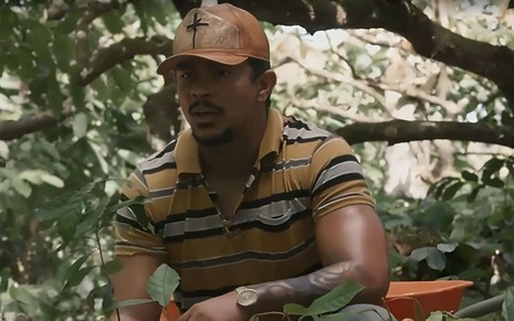 Em cena de Renascer, Xamã usa boné e camisa listrada; ele está agachado em meio à plantação de cacau, falando com alguém