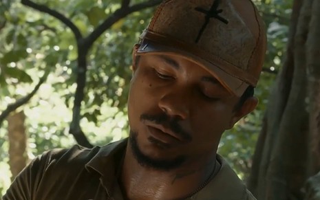 Em cena de Renascer, Xamã usa boné e camisa marrom; ele está trabalhando na plantação de cacau, falando com alguém
