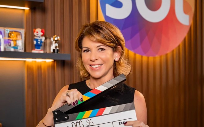 Daniela Beyruti segura claquete em frente a cenário com logo do SBT; ela usa uma blusa preta e sorri para a câmera