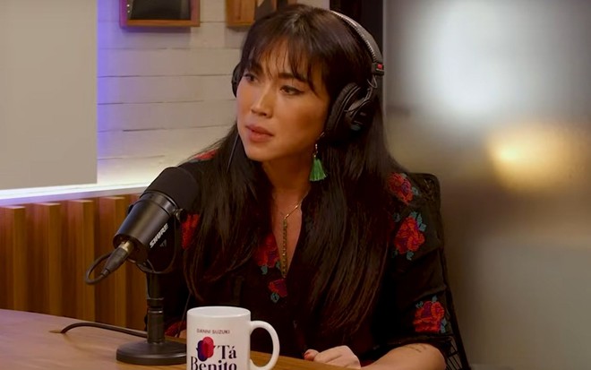 Com fone no ouvido e falando a um microfone, Danni Suzuki está dando entrevista ao podcast Tá Benito