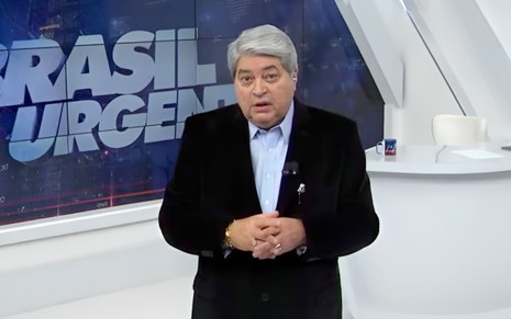 O apresentador José Luiz Datena de terno preto e camisa azul à frente do Brasil Urgente