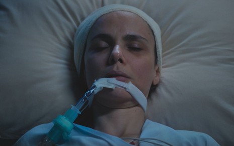 Débora Falabella com olhos fechados e deitada em cama de hospital em cena da novela Terra e Paixão