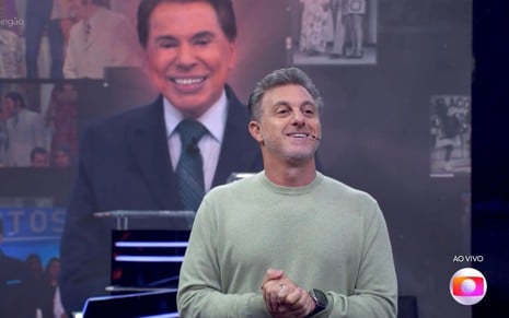 Luciano Huck no palco do Domingão; imagem de Silvio Santos aparece no telão ao fundo