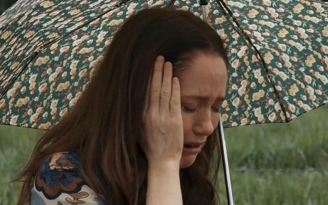 Em cena de Renascer, Camila Morgado chora com a mão no rosto; ela segura um guarda-chuva