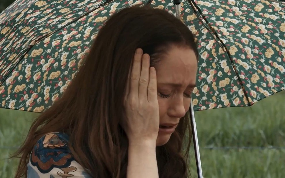 Em cena de Renascer, Camila Morgado chora com a mão no rosto; ela segura um guarda-chuva