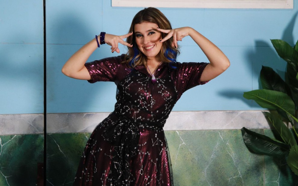Com um vestido lilás, Nicole Orsini faz pose com as mãos próximas ao rosto; ela sorri e tem mechas azuis no cabelo