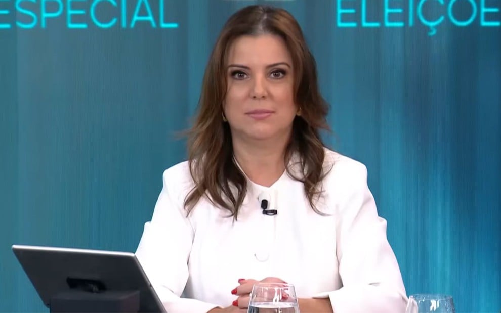 Elaine Keller veste um terno branco e está sentada de braços cruzados com um notebook em cima da mesa, no programa Linha de Frente, da Jovem Pan